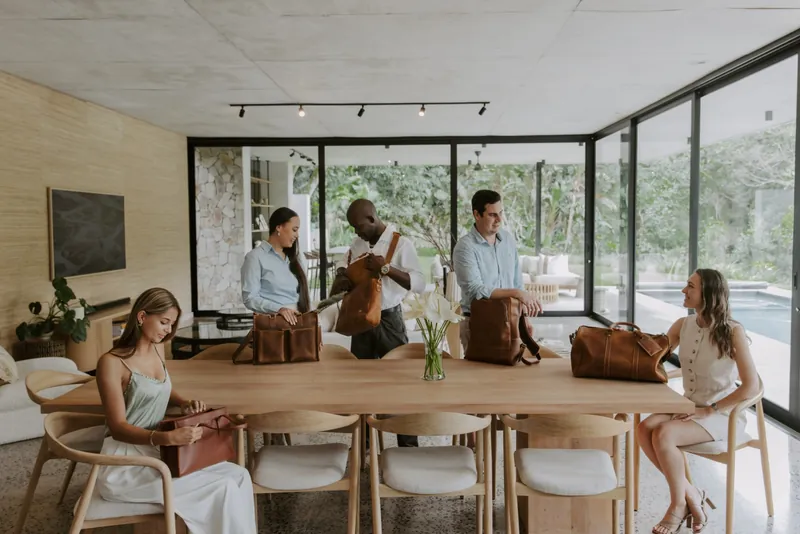 People gathered around a dining table in a bright modern home with Balvaro leather totes, backpacks, and duffels