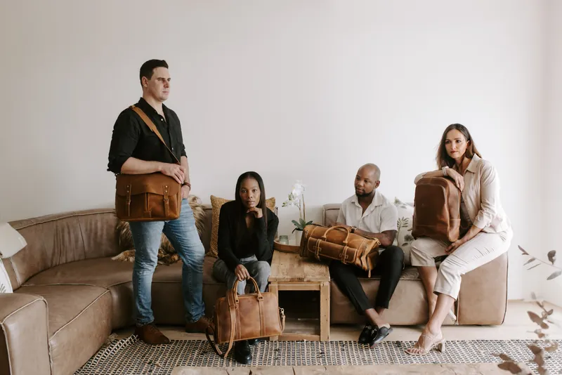 Four people in a living room with Balvaro leather bags: a messenger bag, a laptop bag, a duffel, and a backpack