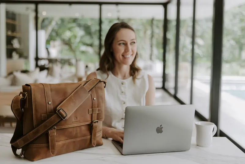 A person working on a laptop at a marble table beside a Balvaro leather messenger bag