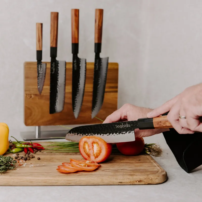 Slicing a tomato with a hammered-finish Soshida santoku, with matching knives on a magnetic wooden block behind